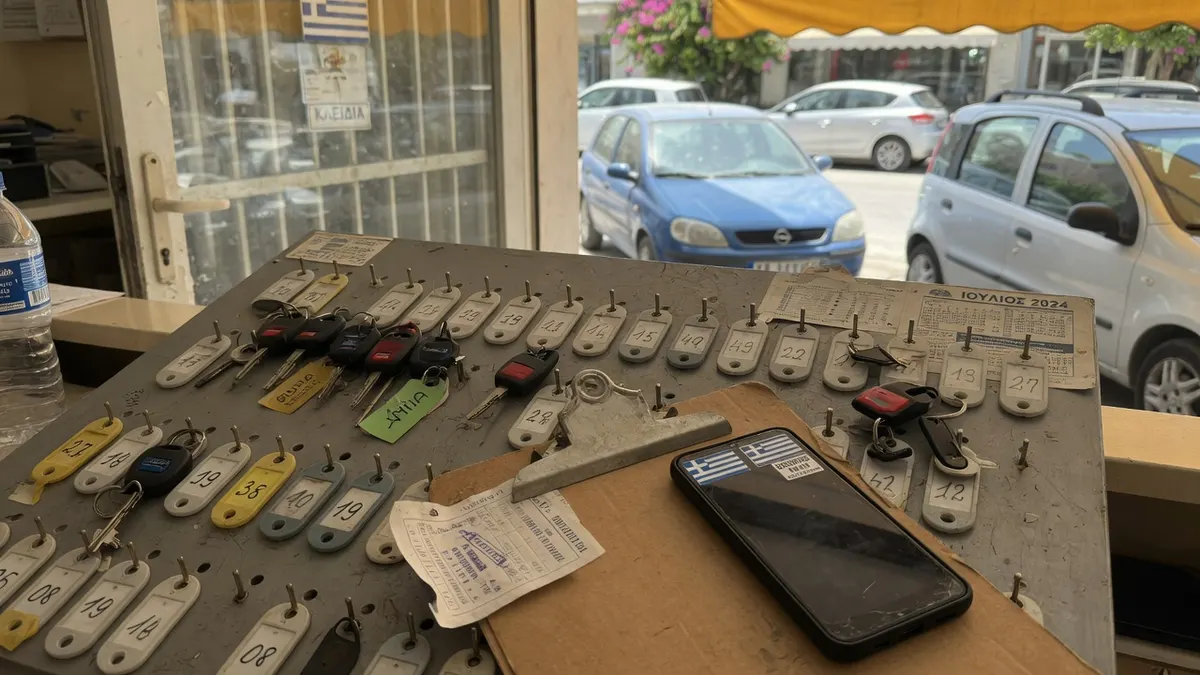 Reihe kleiner Mietwagen auf einem staubigen Platz an einer griechischen Flughafenstraße, Schlüssel am Haken.